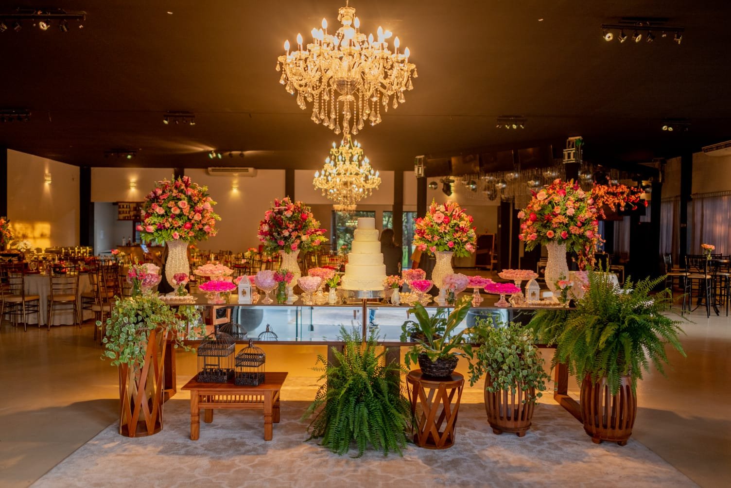 Salão de casamento decorado com mesa do bolo, arranjos florais e lustres de cristal